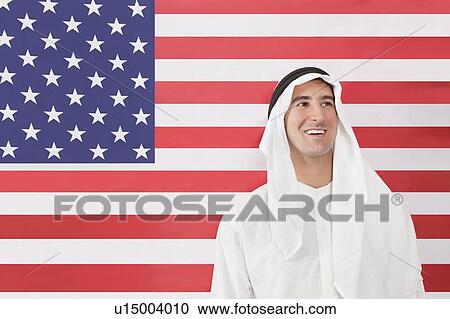 Stock Image - An Arab man in traditional clothes looking away against American flag. Fotosearch