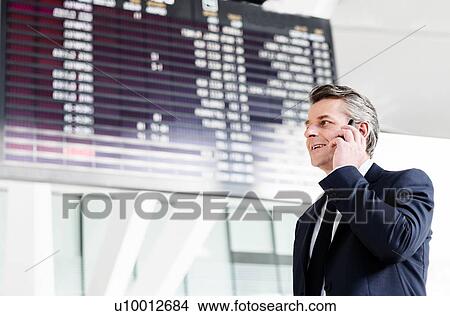Attractive mature businessman talking on smartphone while standing against flight screen display in airport View Large Photo Image Picture - Attractive mature businessman talking on smartphone while standing against flight screen display in airport. Fotosearch