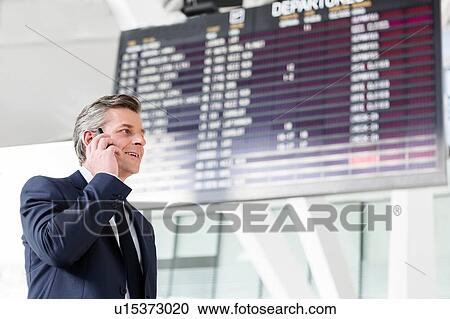Stock Image - Attractive mature businessman talking on smartphone while standing against flight screen display in airport. Fotosearch