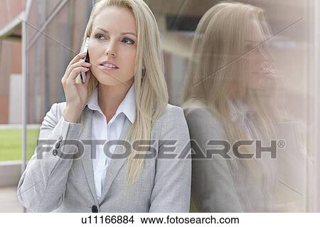 Beautiful blond businesswoman conversing on cell phone while looking away by glass wall View Large Photo Image Picture - Beautiful blond businesswoman conversing on cell phone while looking away by glass wall. Fotosearch