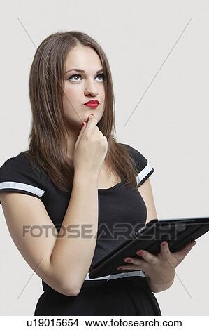 Picture - Beautiful young woman holding tablet PC while looking up over gray background. Fotosearch