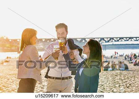 Stock Photo - Business people celebrating success while drinking beer on the beach with lens flare in background. Fotosearch