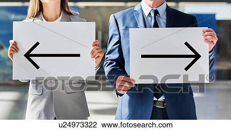 Stock Image - Business people standing while holding white Left and Right arrow placard in arrival area at airport . Fotosearch