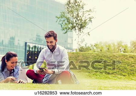 Stock Image - Business people using smartphone while sitting on the grass. Fotosearch
