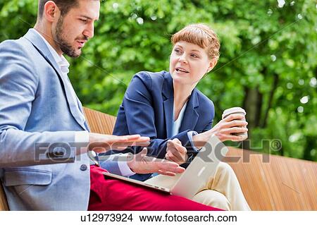 Picture - Businessman discussing plans over laptop with beautiful businesswoman while sitting on bench. Fotosearch