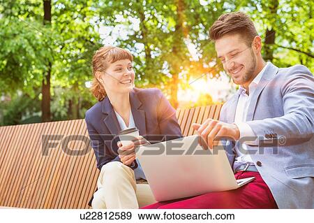 Stock Photo - Businessman discussing plans over laptop with beautiful businesswoman while sitting on bench. Fotosearch