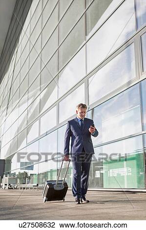 Businessman using smartphone while walking with his suitcase in the airport View Large Photo Image Stock Image - Businessman using smartphone while walking with his suitcase in the airport. Fotosearch