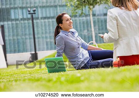 Stock Image - Businesswoman smiling and talking to her colleague while sitting on the grass. Fotosearch