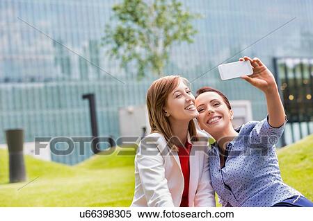 Stock Photography - Businesswomen taking selfie while sitting on the grass in park. Fotosearch