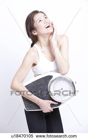 Stock Photography - Cheerful Asian woman with weight scale against white background. Fotosearch
