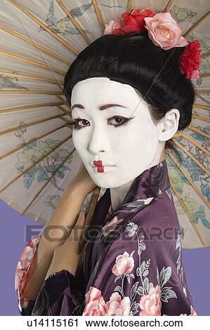 Stock Image - Close-up of Japanese woman with painted face holding parasol against purple background. Fotosearch