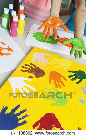 Stock Image - Close-up of paint covered hands and colorful handprints on papers. Fotosearch