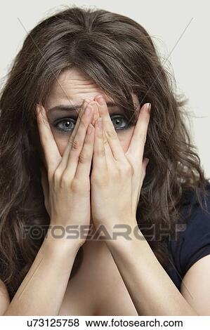 Stock Photo - Close-up portrait of frightened young woman with hands on face against gray background. Fotosearch