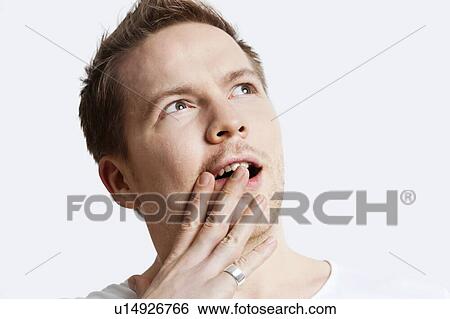 Stock Photograph - Contemplative young man with hand over mouth against white background. Fotosearch
