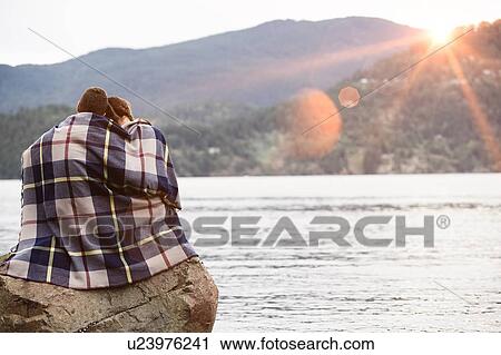 Stock Image - Couple wrapping in blanket sitting by lake. Fotosearch