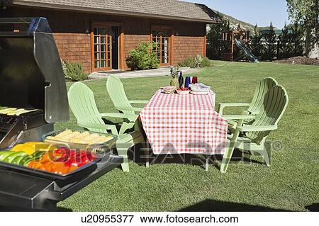 Stock Photo - Dining table and chairs with barbecue grill on lawn in front of country house. Fotosearch