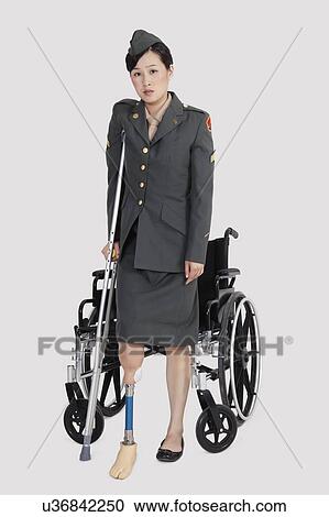 Stock Image - Disabled female US military officer with crutch standing in front of wheelchair over gray background. Fotosearch