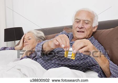 Stock Image - Elderly man tearing condom packet while spouse sleeping behind on bed. Fotosearch
