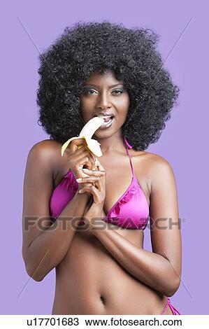 Stock Image - Flirtatious young woman in bikini eating banana against violet background. Fotosearch