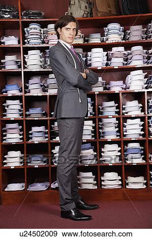 Stock Photo - Full length portrait of confident owner with arms crossed standing against display cabinet. Fotosearch