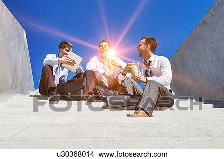 Picture - Group of handsome businessman sitting on stairs while talking during break . Fotosearch
