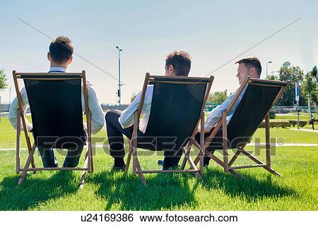 Stock Photograph - Group of handsome businessmen relaxing and talking about life while sitting on folding chair at park. Fotosearch