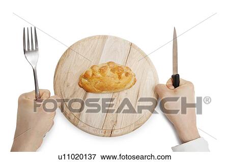 Stock Photo - Hands holding knife and fork with Cornish pastry on wooden plate over white background. Fotosearch