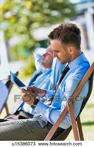 Stock Image - Handsome businessman sitting on folding chair in park while using digital tablet. Fotosearch