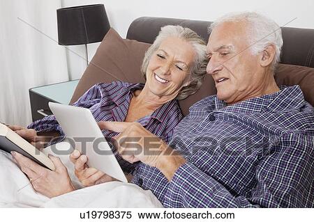 Happy senior couple using digital tablet while lying on bed in room View Large Photo Image Stock Photography - Happy senior couple using digital tablet while lying on bed in room. Fotosearch