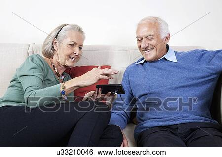 Stock Photograph - Happy senior couple using smart phone while sitting on sofa at home. Fotosearch