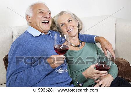 Stock Image - Happy senior couple with red wine glasses while sitting on sofa at home. Fotosearch