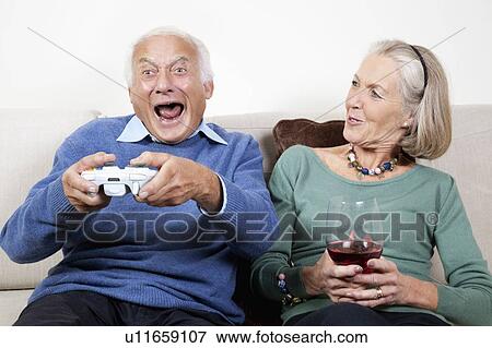 Stock Photo - Happy senior woman with wine glass looking at excited spouse playing video game. Fotosearch
