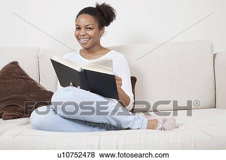 Stock Photo - Happy young African American woman reading book on sofa. Fotosearch