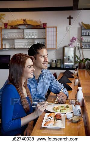 Stock Photo - Happy young couple sitting at counter with sushi while looking away in restaurant. Fotosearch