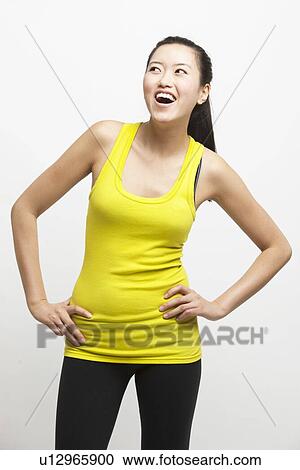 Stock Image - Happy young woman in yellow tank top with hands on hips looking away against white background. Fotosearch