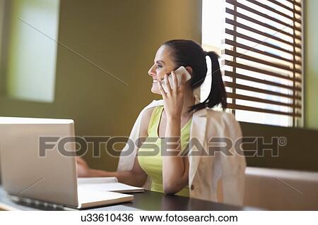 Happy young woman sitting at desk with laptop using cell phone View Large Photo Image Stock Photograph - Happy young woman sitting at desk with laptop using cell phone. Fotosearch