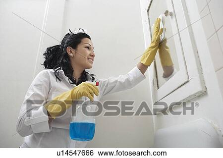 Stock Photograph - Low angle view of young housemaid cleaning mirror. Fotosearch