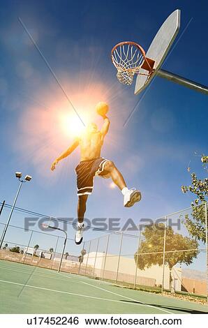 Stock Photograph - Man about to slam dunk ball on basketball court with strong sun shining lensflare. Fotosearch