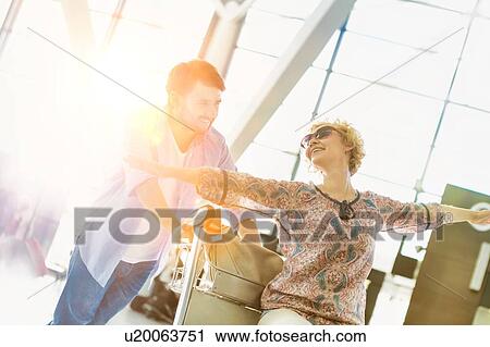 Stock Image - Man pushing baggage cart for check in while his wife is sitting in airport . Fotosearch