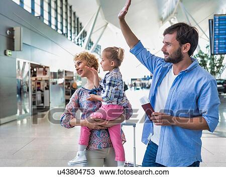 Stock Image - Man waving and holding his passport boarding pass while his wife is carrying their daughter. Fotosearch