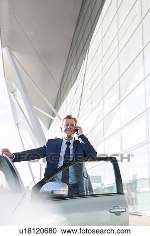 Stock Image - Mature attractive businessman smiling while talking on smartphone outside the car in airport. Fotosearch