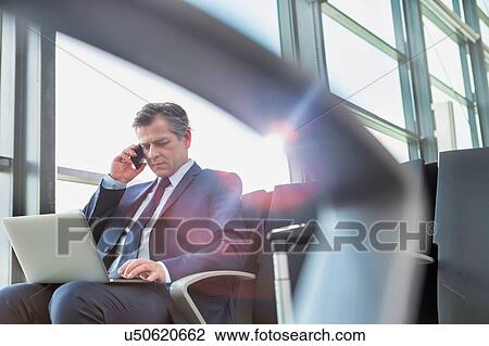 Mature attractive businessman talking on smartphone while sitting and working with his laptop before boarding in airport View Large Photo Image Stock Image - Mature attractive businessman talking on smartphone while sitting and working with his laptop before boarding in airport. Fotosearch