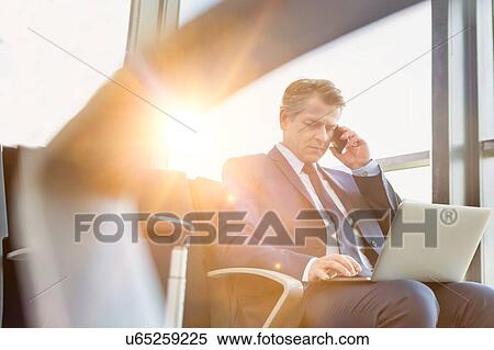 Stock Photography - Mature attractive businessman talking on smartphone while sitting and working with his laptop before boarding in airport. Fotosearch