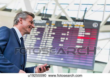 Mature attractive businessman using smartphone while standing against flight display screen in airport View Large Photo Image Stock Image - Mature attractive businessman using smartphone while standing against flight display screen in airport. Fotosearch