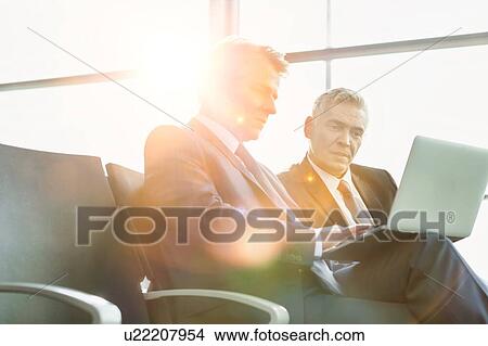 Picture - Mature businessman reading news paper while the other businessman is talking on smartphone in airport. Fotosearch