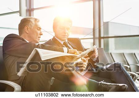 Stock Image - Mature businessman reading news paper while the other businessman is talking on smartphone in airport. Fotosearch