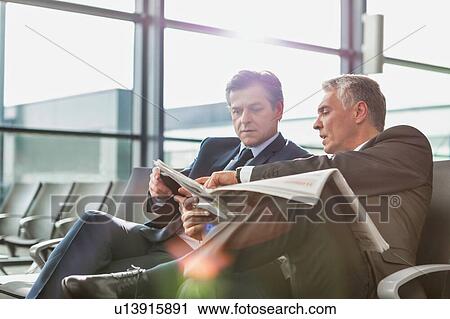 Stock Image - Mature businessman reading news paper while the other businessman is talking on smartphone in airport. Fotosearch