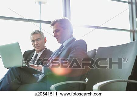 Stock Image - Mature businessman reading news paper while the other businessman is talking on smartphone in airport. Fotosearch