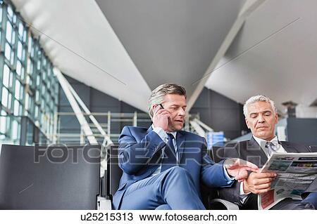 Stock Image - Mature businessman reading news paper while the other businessman is talking on smartphone in airport. Fotosearch