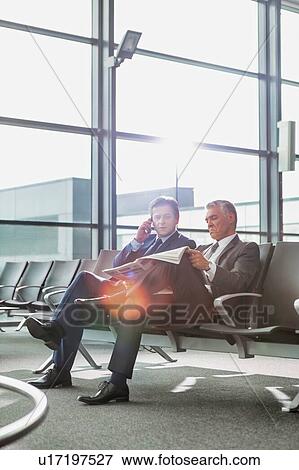 Stock Photo - Mature businessman reading news paper while the other businessman is talking on smartphone in airport. Fotosearch